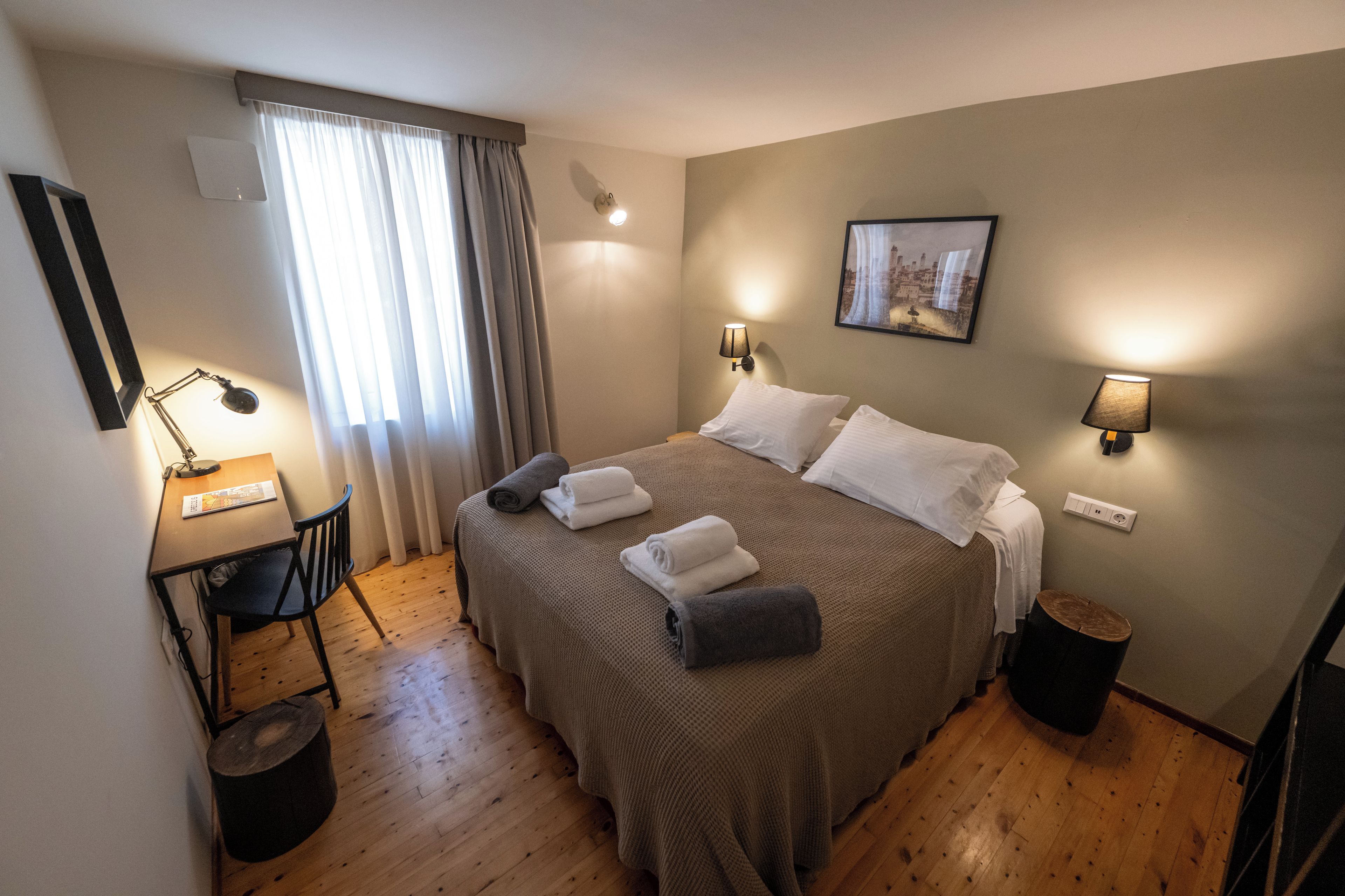 A neatly arranged hotel room with a large bed, bedside lamps, a desk with a chair, and a small trash bin.