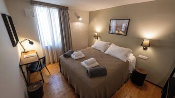 A neatly arranged hotel room with a large bed, bedside lamps, a desk with a chair, and a small trash bin.