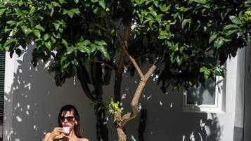 A woman sitting on a white chair under a tree, holding a cup, with a table beside her containing a glass of orange juice, a small plant, and a notebook.