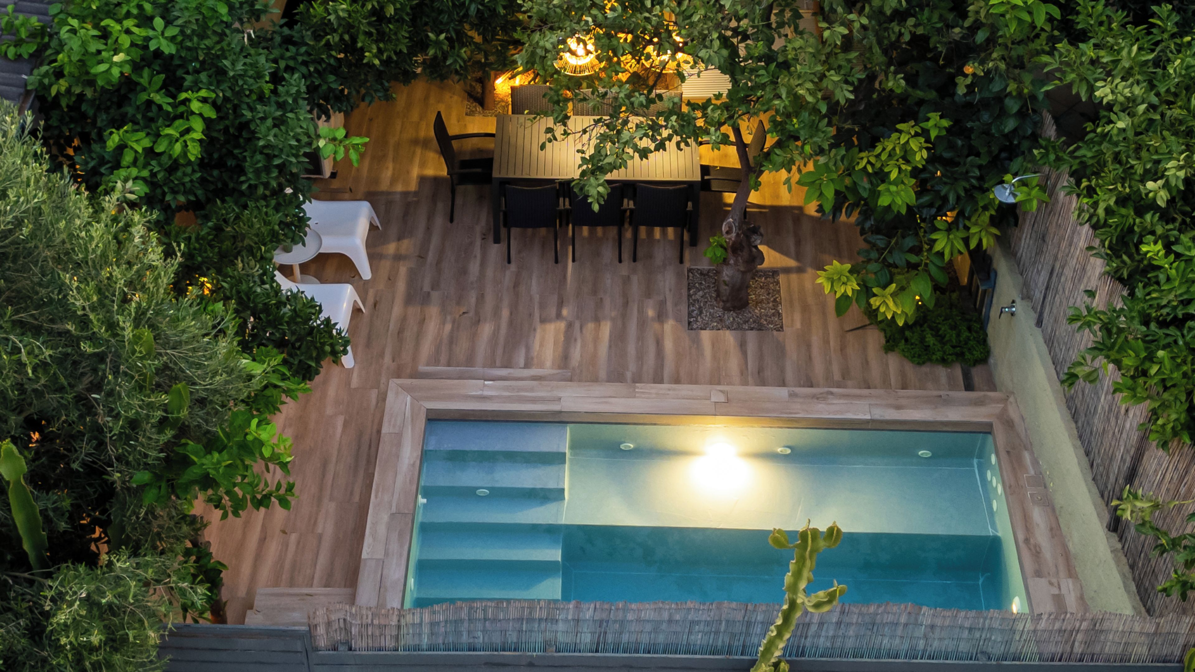An aerial view of a swimming pool surrounded by wooden decking, with a dining area and chairs visible in the background.