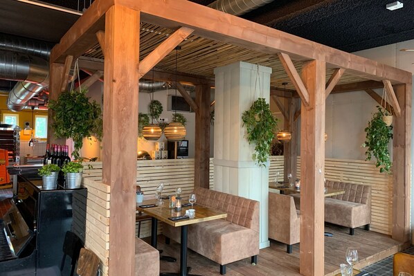 Un restaurante con vigas de madera, asientos acolchados, plantas colgantes y una cocina a la vista.