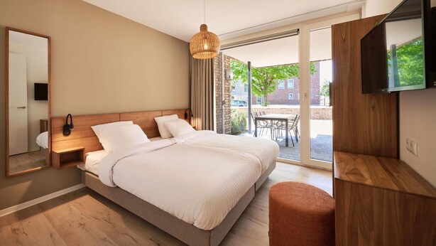 A modern bedroom with a large bed, a TV mounted on the wall, and a sliding glass door leading to an outdoor area. - Castellum Spa Apartment (Maastricht)