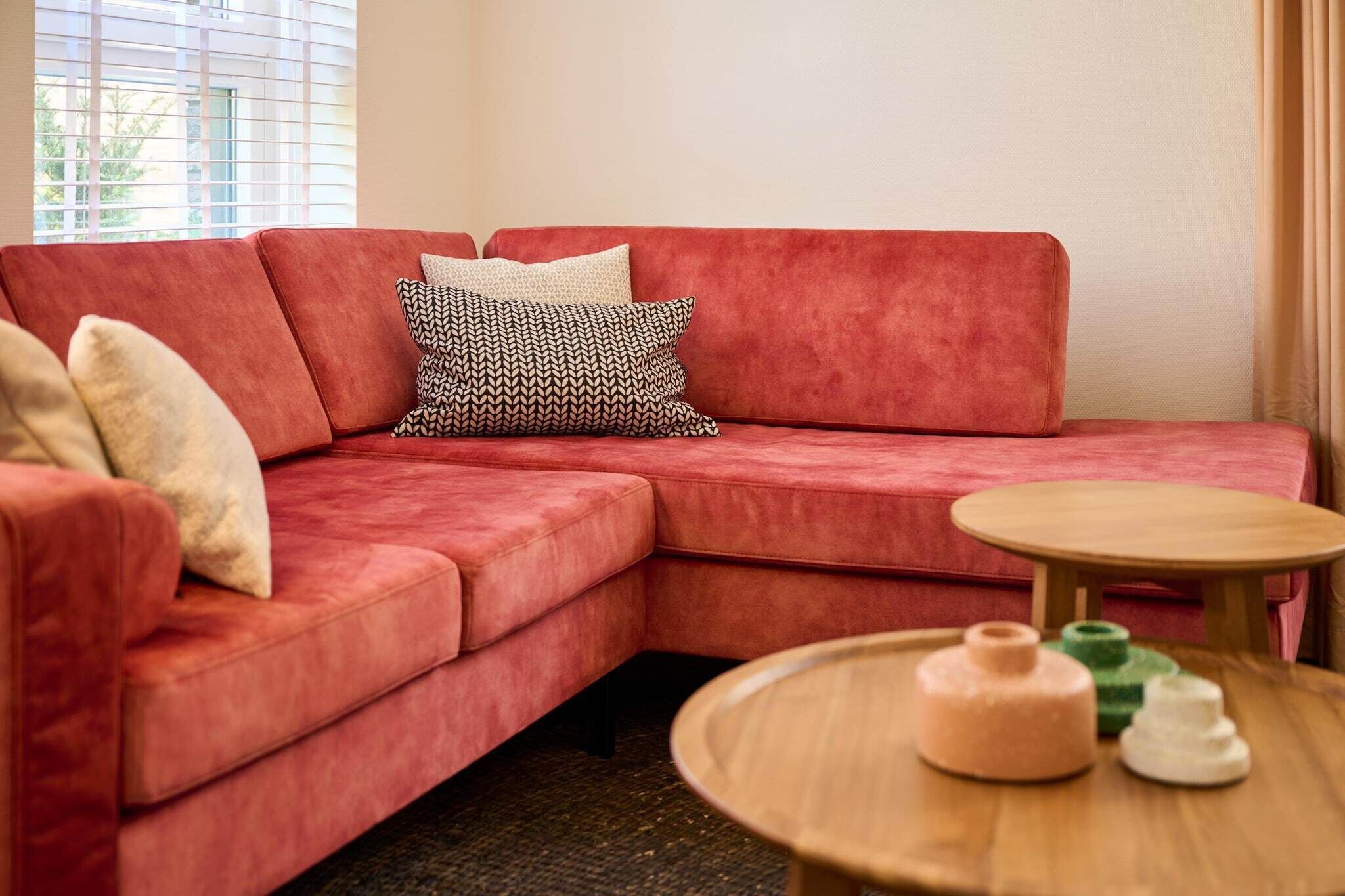 Ein rotes Modulsofa mit gemusterten Kissen, ein kleiner Holztisch und ein Fenster mit Jalousien.