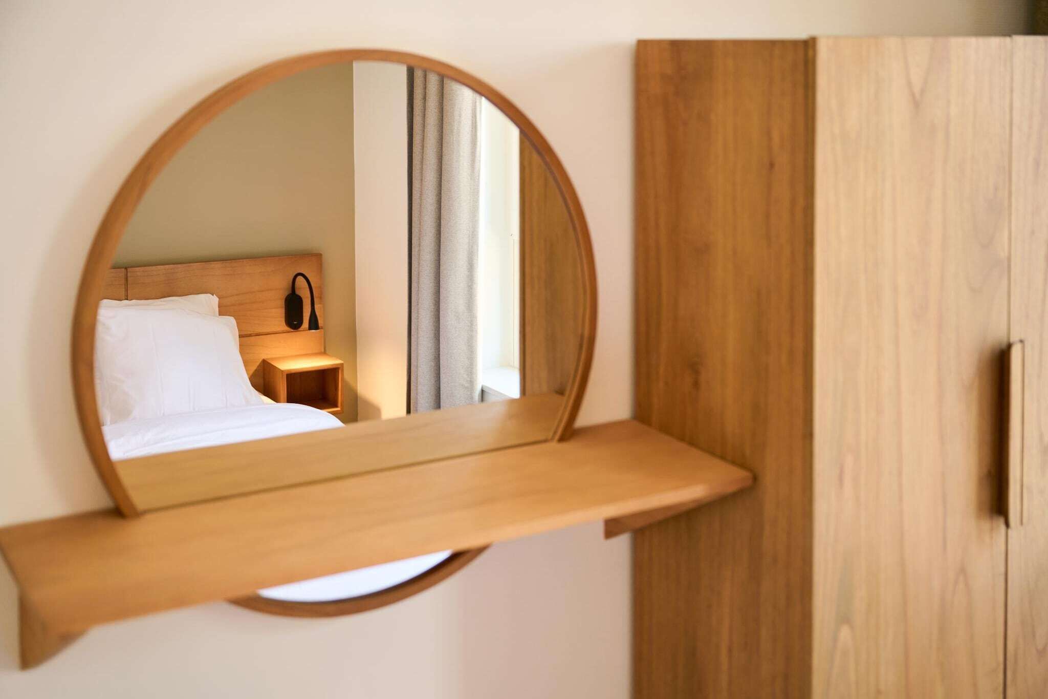 A circular mirror with a wooden frame mounted on a wall, reflecting a bed and a nightstand.