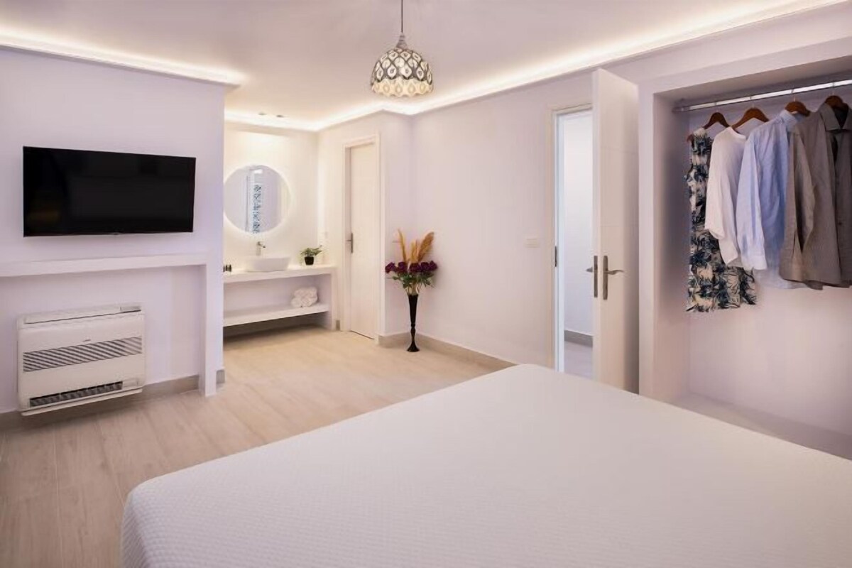 A modern hotel room with a flat-screen TV, a bedside table, a wardrobe, and a bathroom visible through an open door.