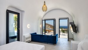 Un dormitorio moderno con cama, sofá azul y televisor. Hay dos puertas que dan a un balcón con vistas al mar.