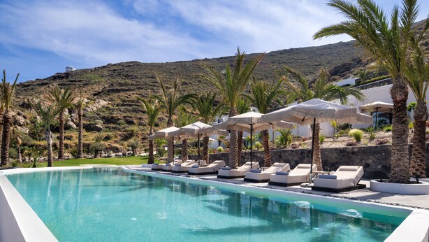 Un área de piscina con sillones reclinables y sombrillas, rodeada de palmeras y montañas al fondo.