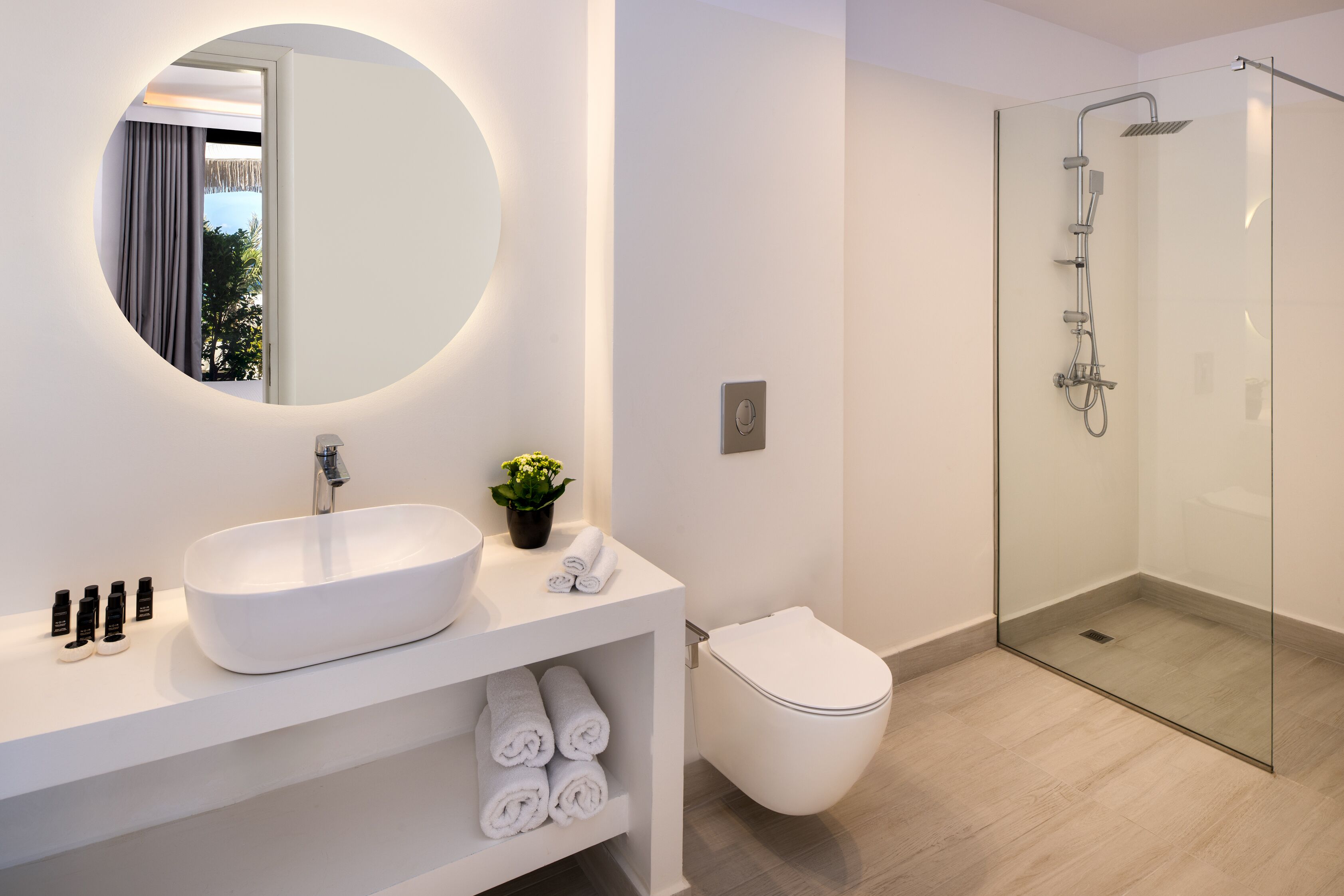 A modern bathroom with a large round mirror, a white sink, a glass-enclosed shower, and a toilet.