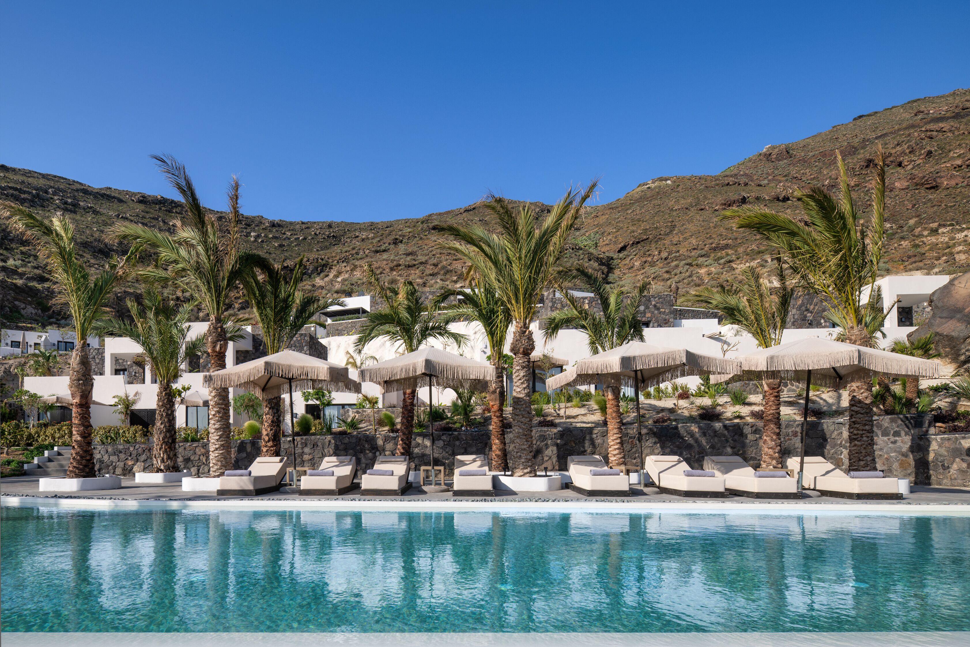 Un espace piscine agrémenté de chaises longues et de parasols, entouré de palmiers et dominé par un paysage montagneux.
