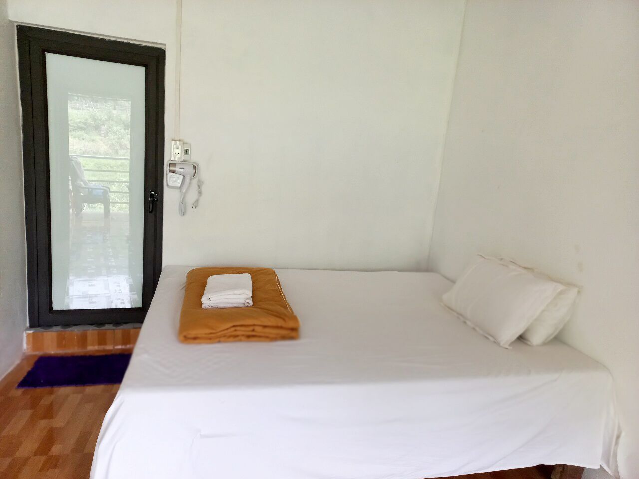 Una cama pequeña y ordenada con sábanas blancas, una toalla naranja doblada y un almohadón blanco.