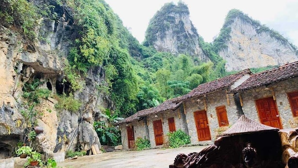 Um edifício de pedra tradicional com portas vermelhas e telhado em cerâmica, rodeado por vegetação exuberante e falésias rochosas.