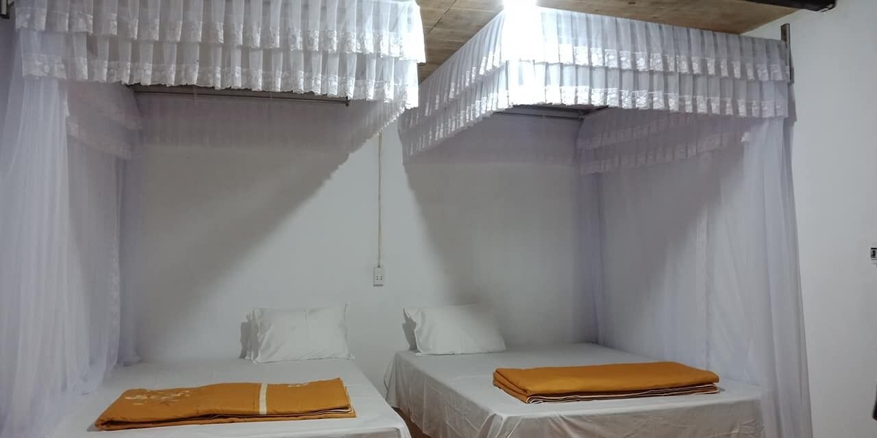 A room with two single beds, each with a white bedspread and a brown blanket. The room has white curtains and a wooden ceiling.