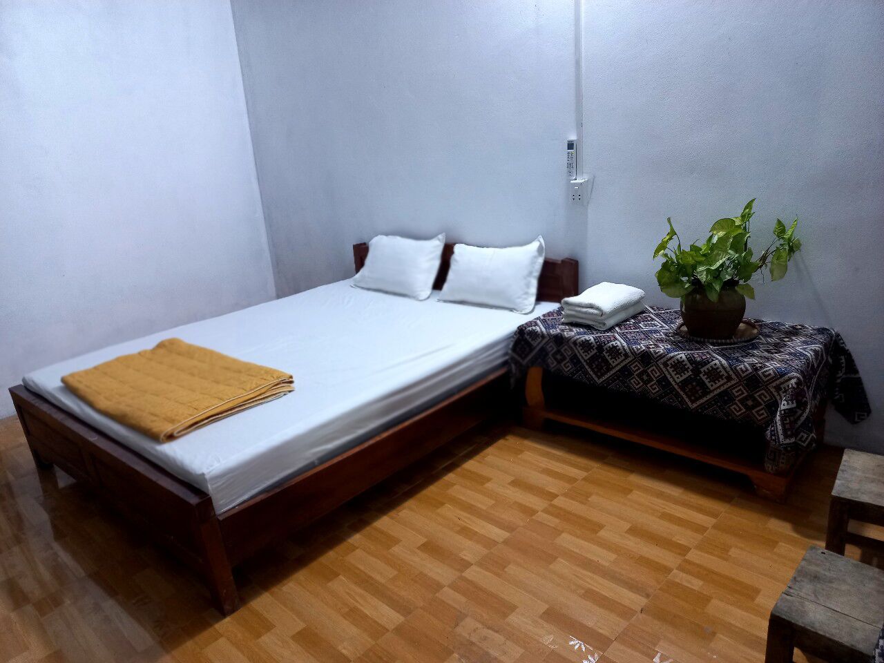 Una cama individual con colchón blanco y manta marrón, una mesita de noche con una planta, y suelo de madera.