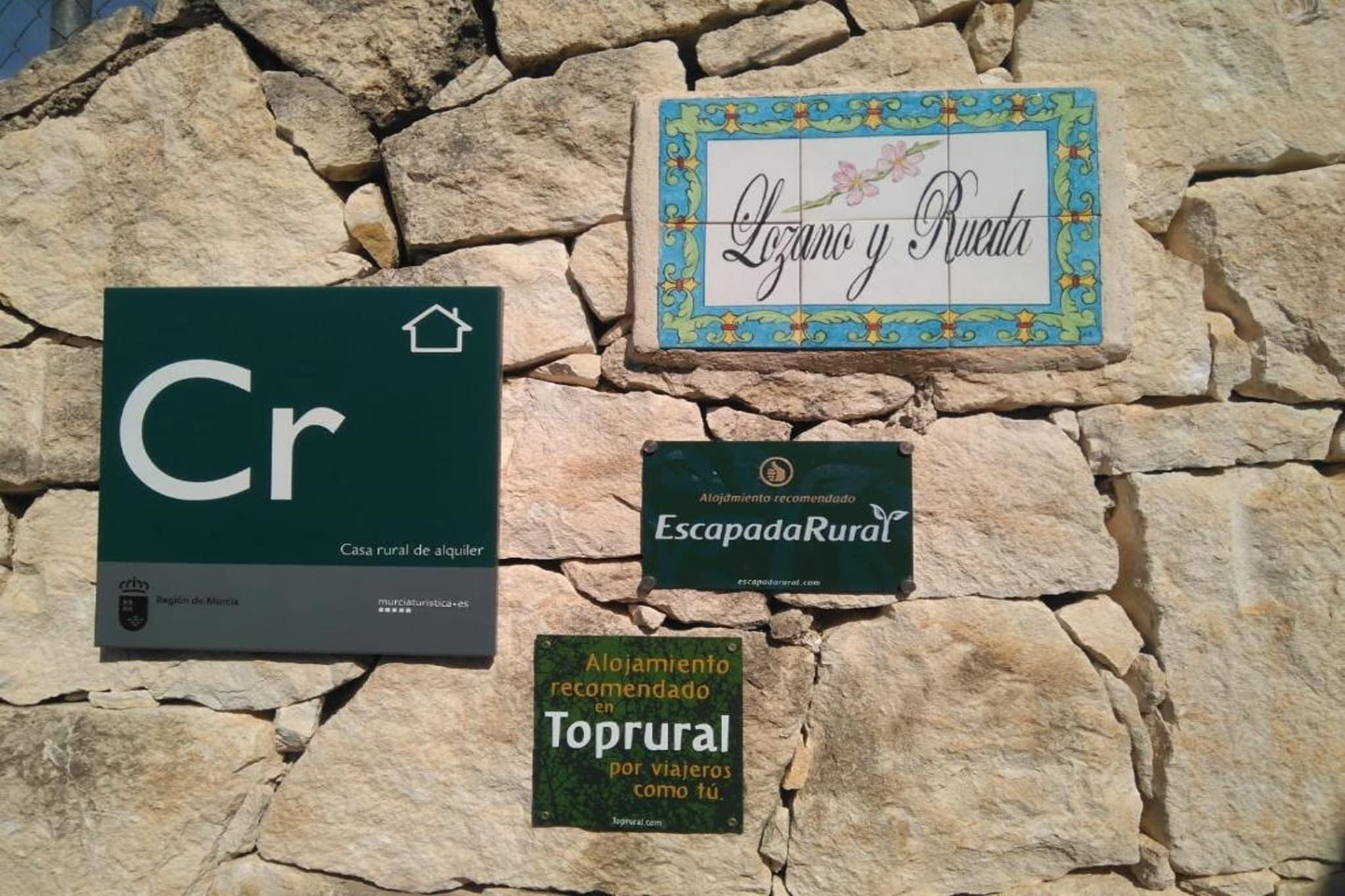Tres señales en un muro de piedra: una señal verde con 'Cr' y 'Casa rural de alquería', una señal decorativa con 'Lozano y Rueda', y una señal verde que recomienda alojamiento 'Toprural'.