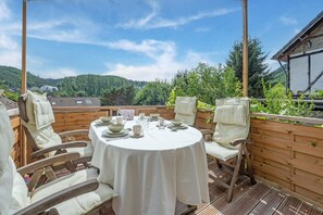 A wooden deck with a round table set for dining, surrounded by cushioned chairs, overlooking a forested hillside. - Pretty Holiday Home in Schleiden with Balcony and Barbecue (Schleiden)