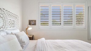 A bedroom with a bed, a nightstand with a lamp, a decorative wall panel, and a window with shutters. - Sea Haven - Lennox Head (Lennox Head)