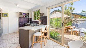 A modern kitchen with a central island, bar stools, and a sliding glass door leading to an outdoor patio with a table and chairs. - Sea Haven - Lennox Head (Lennox Head)