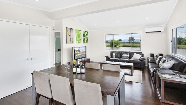 Uma sala ampla com uma mesa de jantar, sofás e uma televisão. Há garrafas de vinho na mesa e uma vista panorâmica pelas janelas.