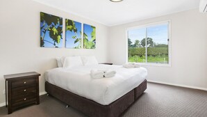 Cama bem arrumada com lençóis brancos e duas almofadas, uma pequena mesa de cabeceira com gavetas, e uma janela grande com vista para o verde lá fora.
