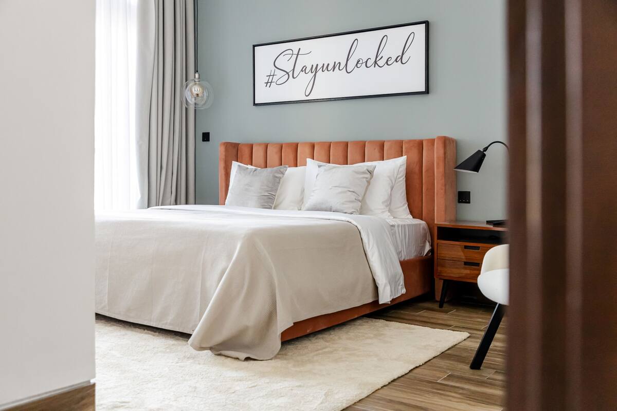 A modern bedroom with a large bed, a bedside table, a lamp, and a wall-mounted sign.