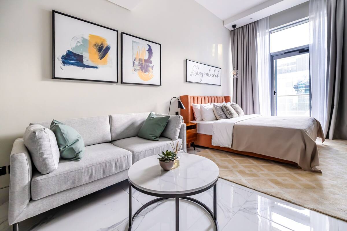 A modern hotel room with a grey sofa, a round coffee table with a plant, a bed with a beige bedspread, and artwork on the walls.