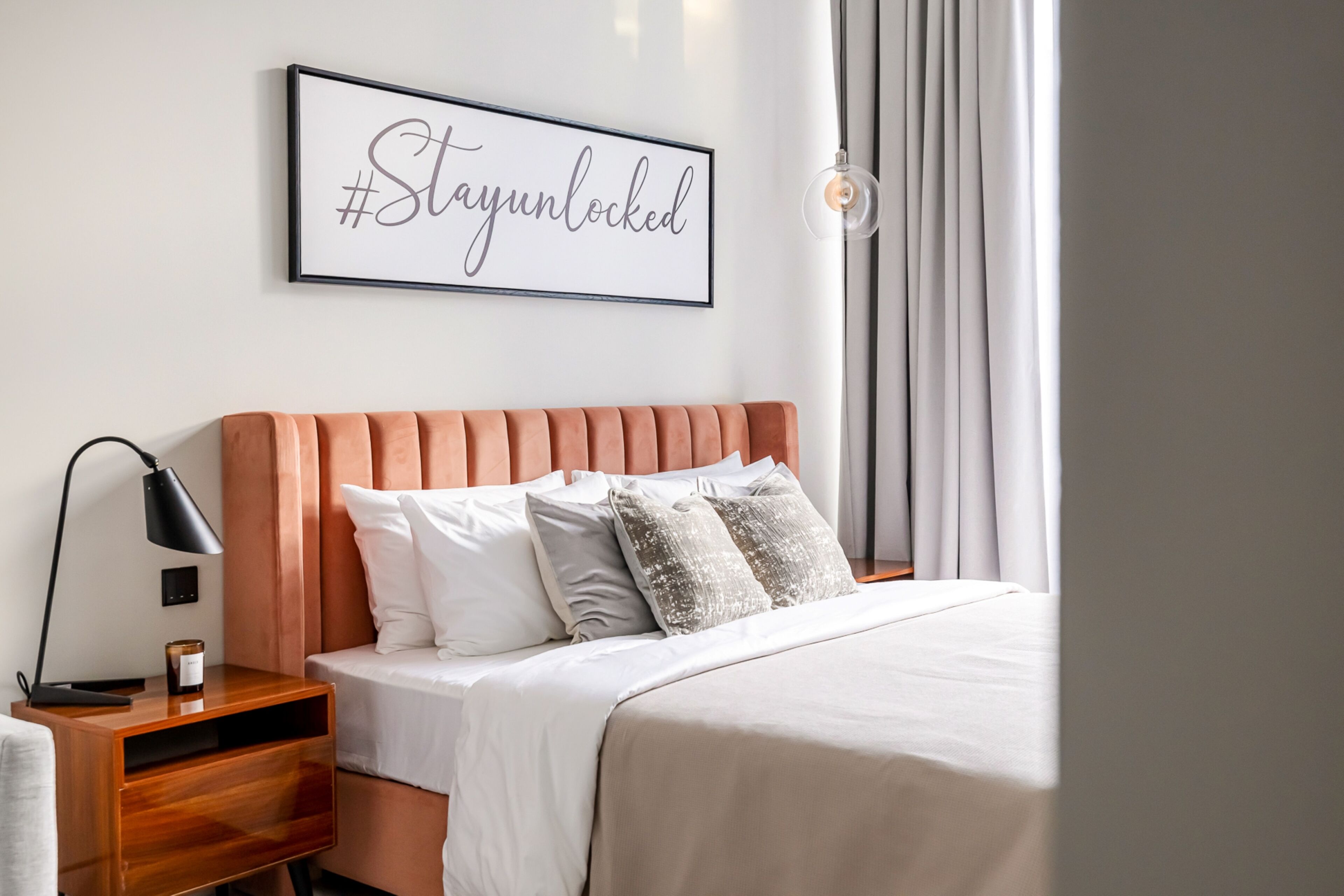 Een netjes opgemaakt bed met hoofdbord, een nachtkastje met lamp, en een lijst aan de muur.
