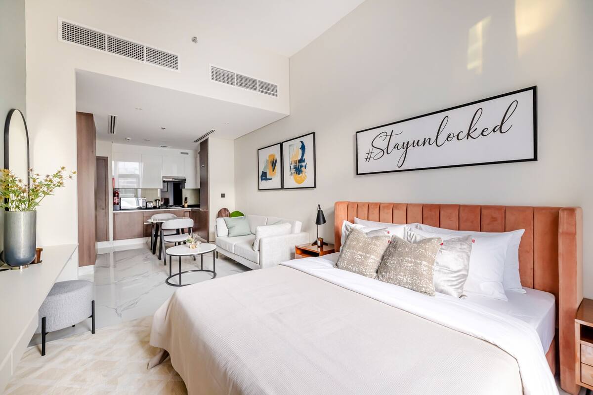 A modern hotel room with a large bed, a sofa, a dining area, and a kitchen in the background.