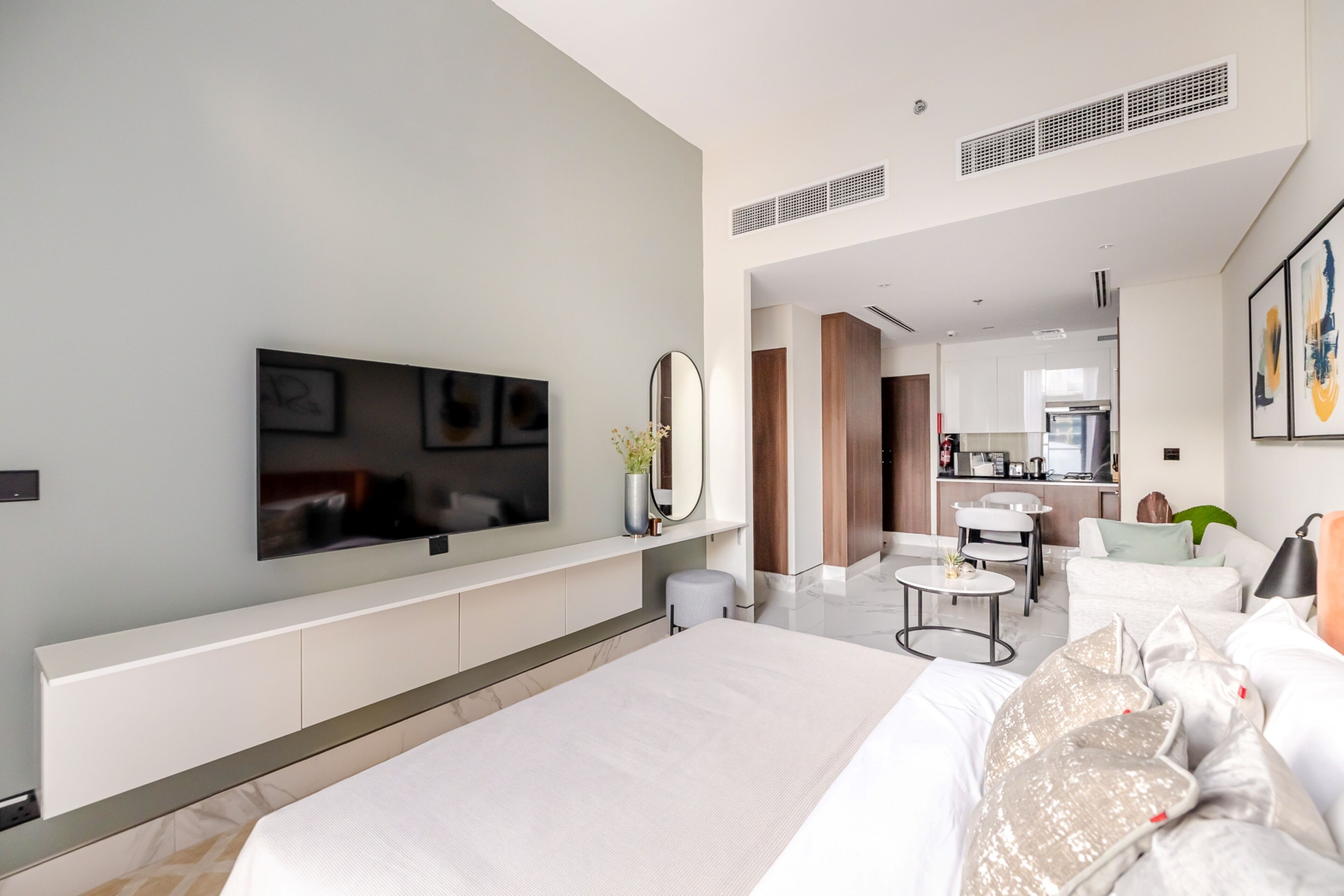 A modern hotel room with a large bed, a flat-screen TV, a mirror, and a small kitchenette in the background.