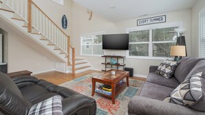 A living room with a staircase, a flat-screen TV, and a 'Every Summer Has a Story' sign. - 5136- Bayside Breeze - Soundside KDH Retreat | Dog-Friendly (Kill Devil Hills)