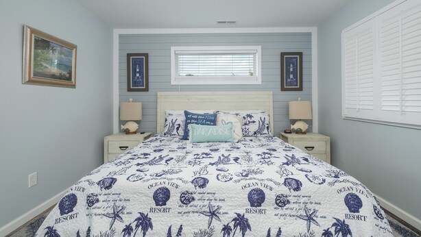 A bedroom with a bed, nightstands, a window with blinds, and framed pictures on the wall. - 5136- Bayside Breeze - Soundside KDH Retreat | Dog-Friendly (Kill Devil Hills)