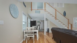 A spacious living area with a dining table, chairs, a brown leather couch, and a staircase leading to an upper level. - 5136- Bayside Breeze - Soundside KDH Retreat | Dog-Friendly (Kill Devil Hills)
