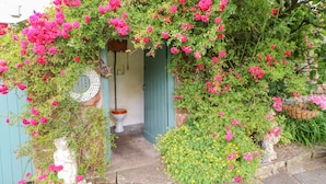 A rustic outdoor entrance with a stone pathway leading to a wooden door, surrounded by lush greenery and vibrant pink flowers. - Eden Cottage (Appleby-in-Westmorland)