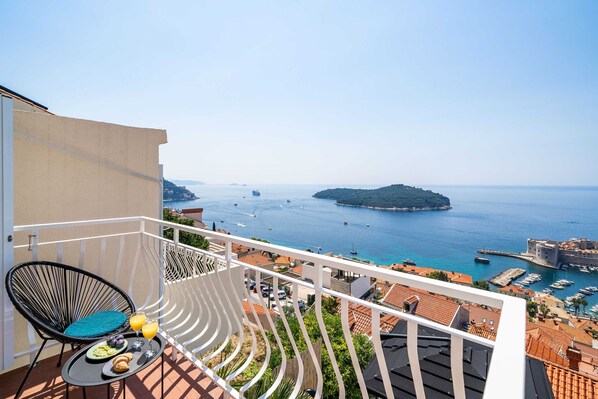 A balcony with a view of the sea, a chair, a table with food, and a cityscape in the background.