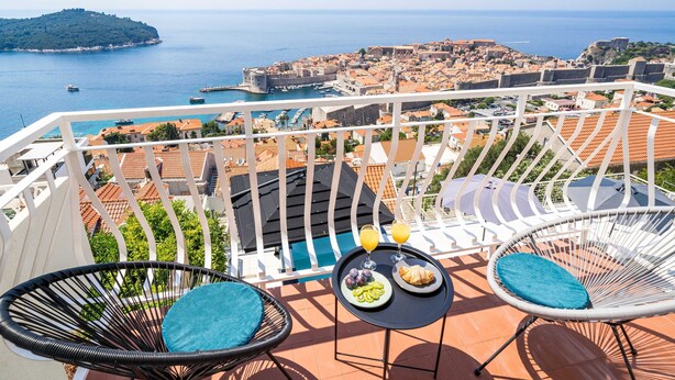 A balcony with two wicker chairs, a small table with breakfast, and a view of a coastal town and sea. - Apartment Raguseo (Dubrovnik)