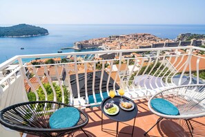 A balcony with two wicker chairs, a small table with breakfast, and a view of a coastal town and sea.
