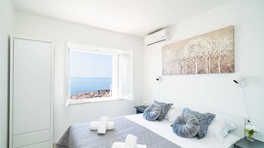 A bedroom with a bed, pillows, a window with a view, and a painting on the wall. - Apartment Raguseo (Dubrovnik)