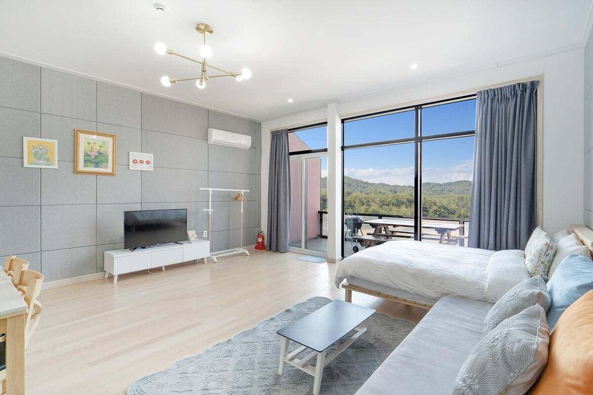 A modern hotel room with a large bed, a sofa, a TV, and a balcony with a scenic view.