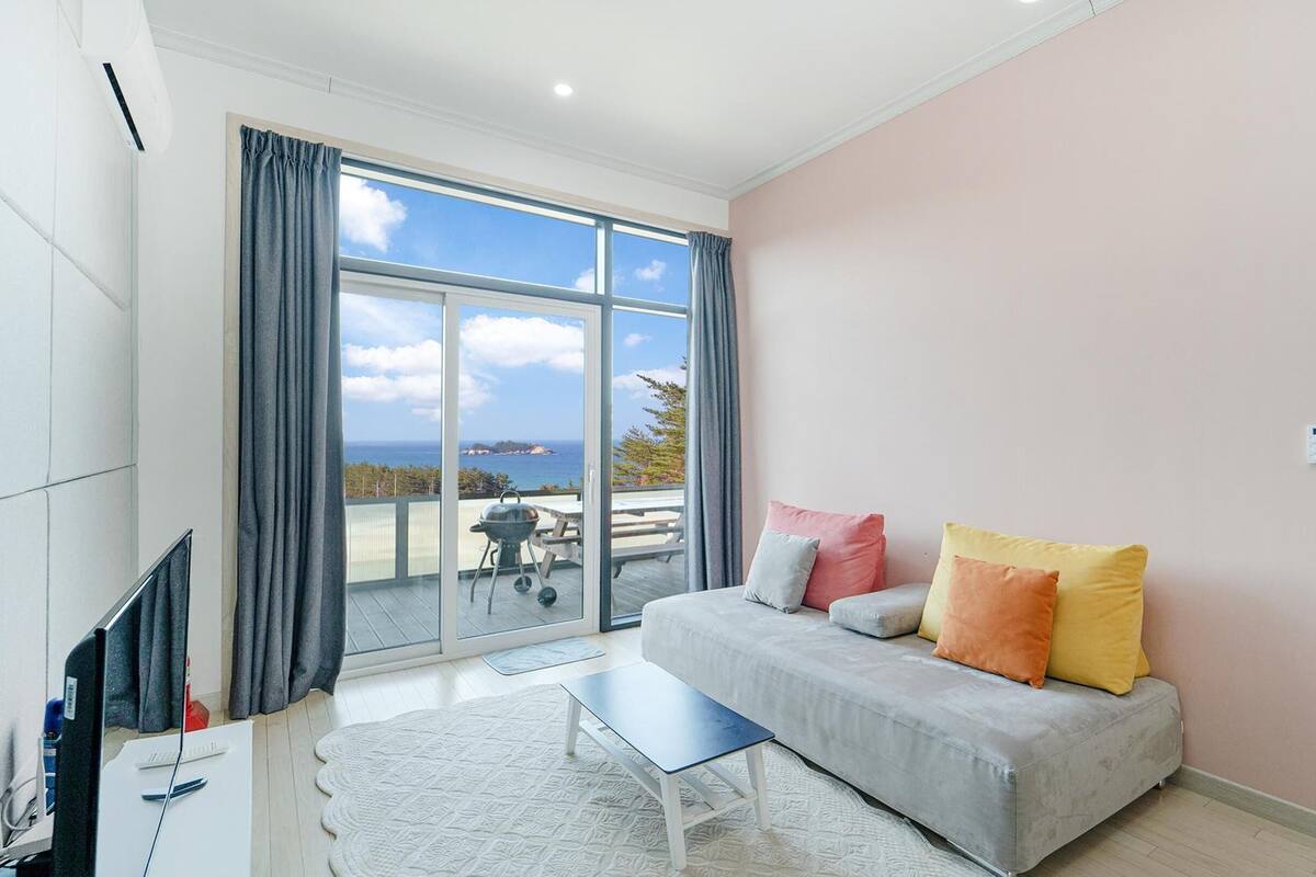 A modern living room with a grey sofa, a small white table, and a view of the sea through large windows.