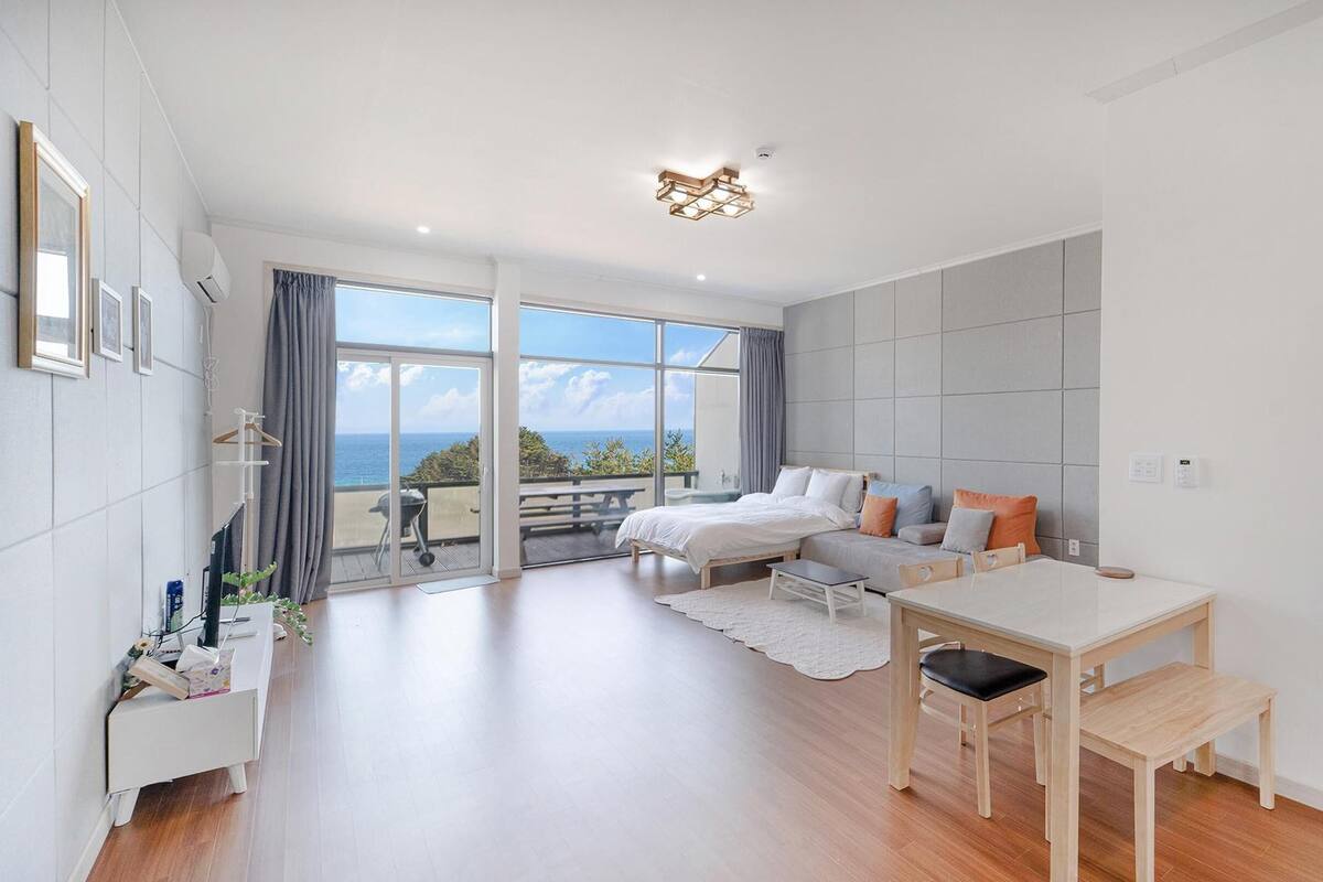 A modern bedroom with a large window, a bed, a sofa, a dining table, and a view of the ocean.