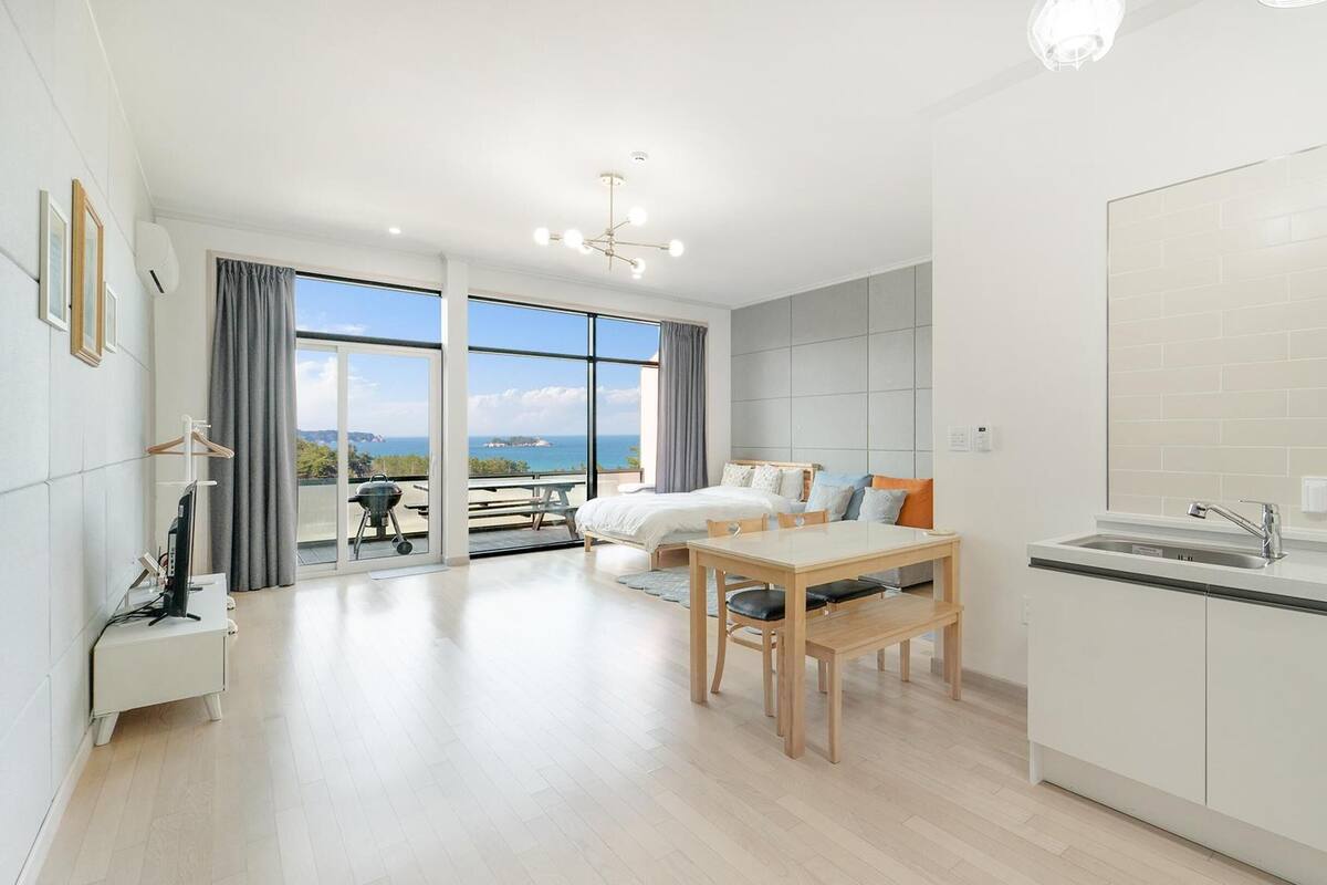 A modern hotel room with a balcony, a kitchenette, and a large window offering a sea view.