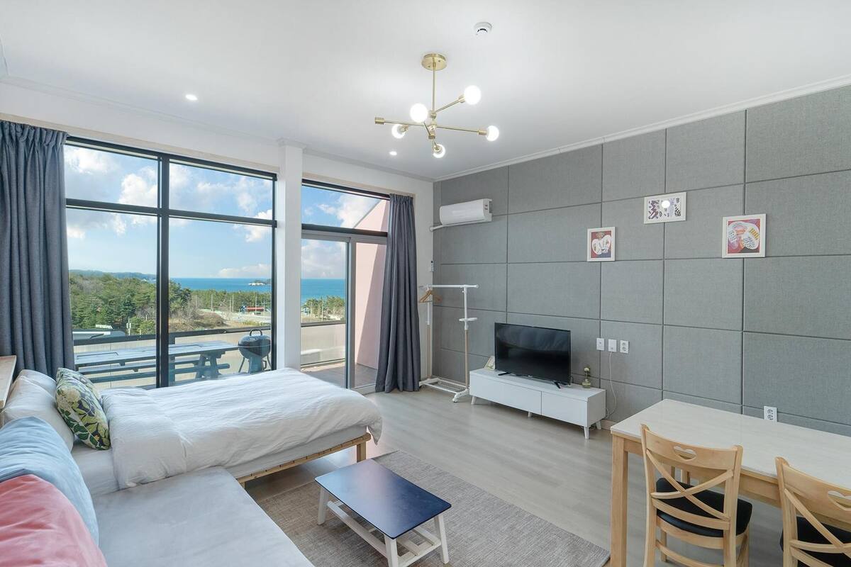 A modern hotel room with a large bed, a TV, a dining area, and a balcony with a view of the sea.