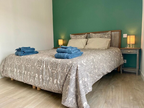 Ein ordentlich bezogenes Bett mit einem gemusterten Überwurf, zwei Kissen und gefalteten Handtüchern an der Seite. Ein Nachttisch mit Lampe und kleiner Schublade steht daneben. Der Raum hat Holzböden und grüne Wände.