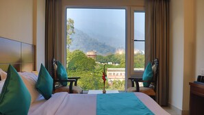 A hotel room with a large bed, two chairs, a small table, and a view of a mountainous landscape through a large window. - Hotel Green - Behind Parmarth Niketan (Rishikesh)