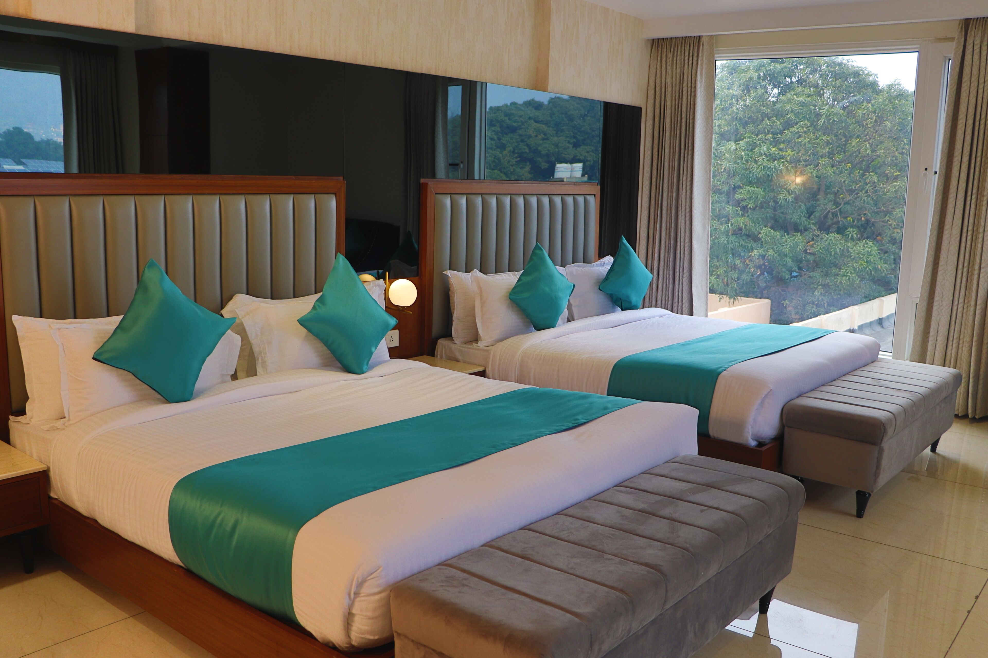 A hotel room with two beds, each with a headboard, a bench, and a nightstand with a lamp.