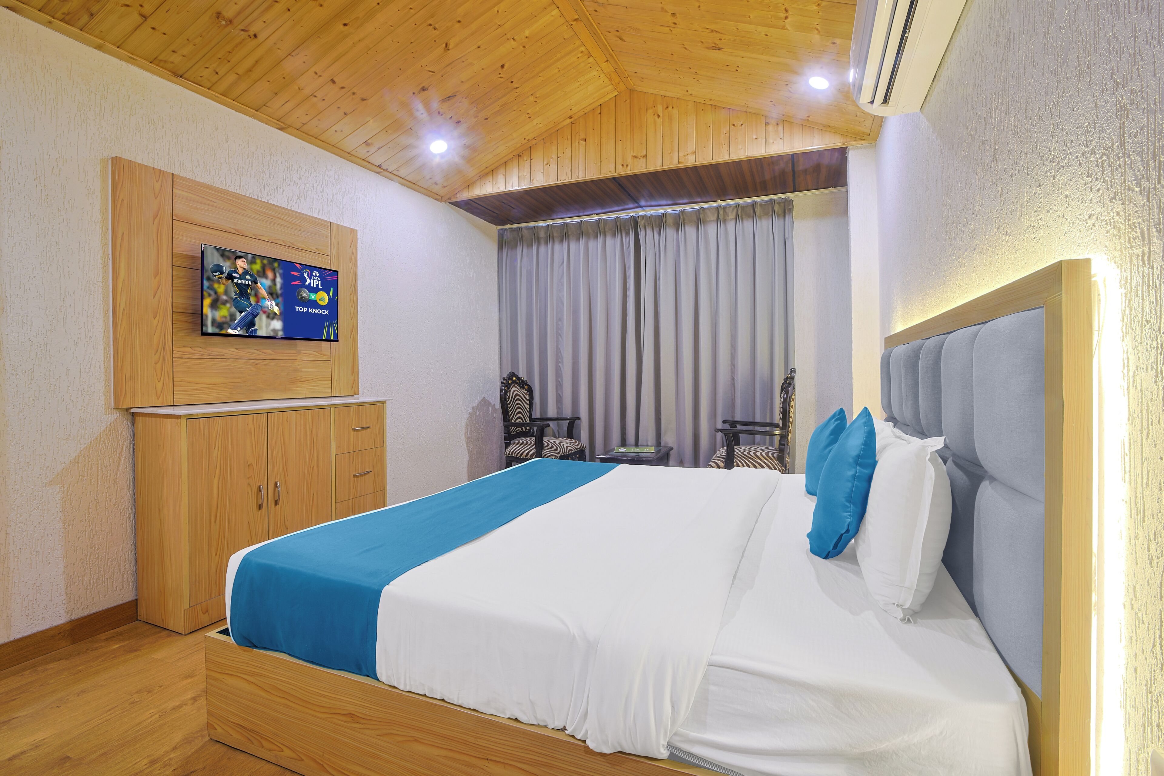 Une chambre d’hôtel avec un lit, un téléviseur fixé au mur, un plafond en bois et une fenêtre avec des rideaux.