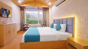 A hotel room with a wooden ceiling, a large bed with blue and white bedding, a TV on the wall, and a view of mountains through the window.
