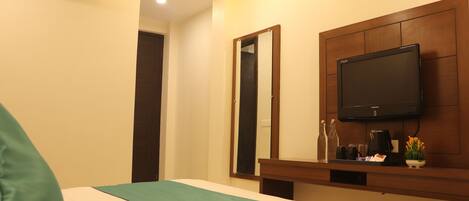 A hotel room with a bed, a mounted television, a mirror, a wooden headboard, and a small table with a coffee maker and a plant.