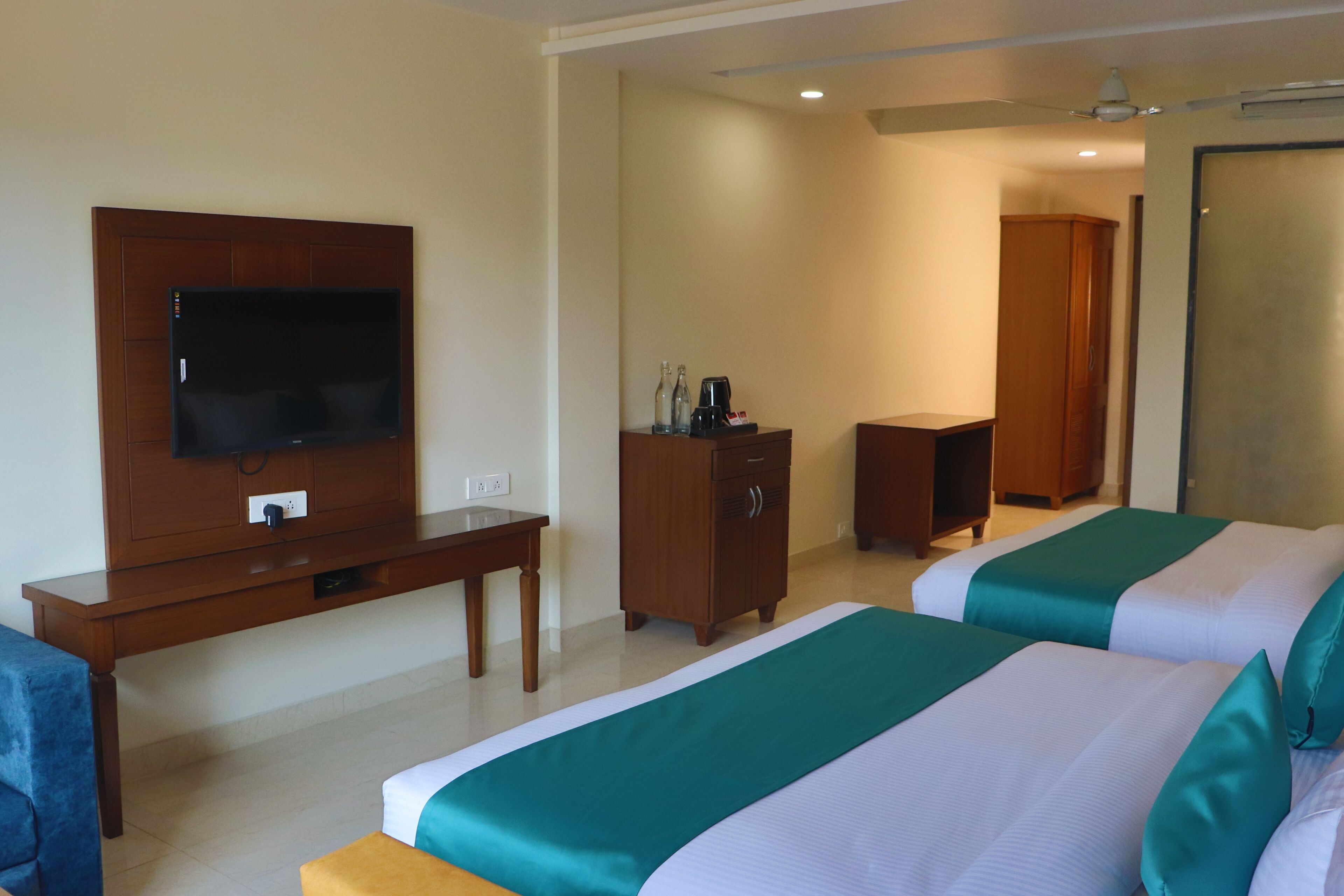 A hotel room with two beds, a TV, a wooden desk, a wardrobe, and a ceiling fan.