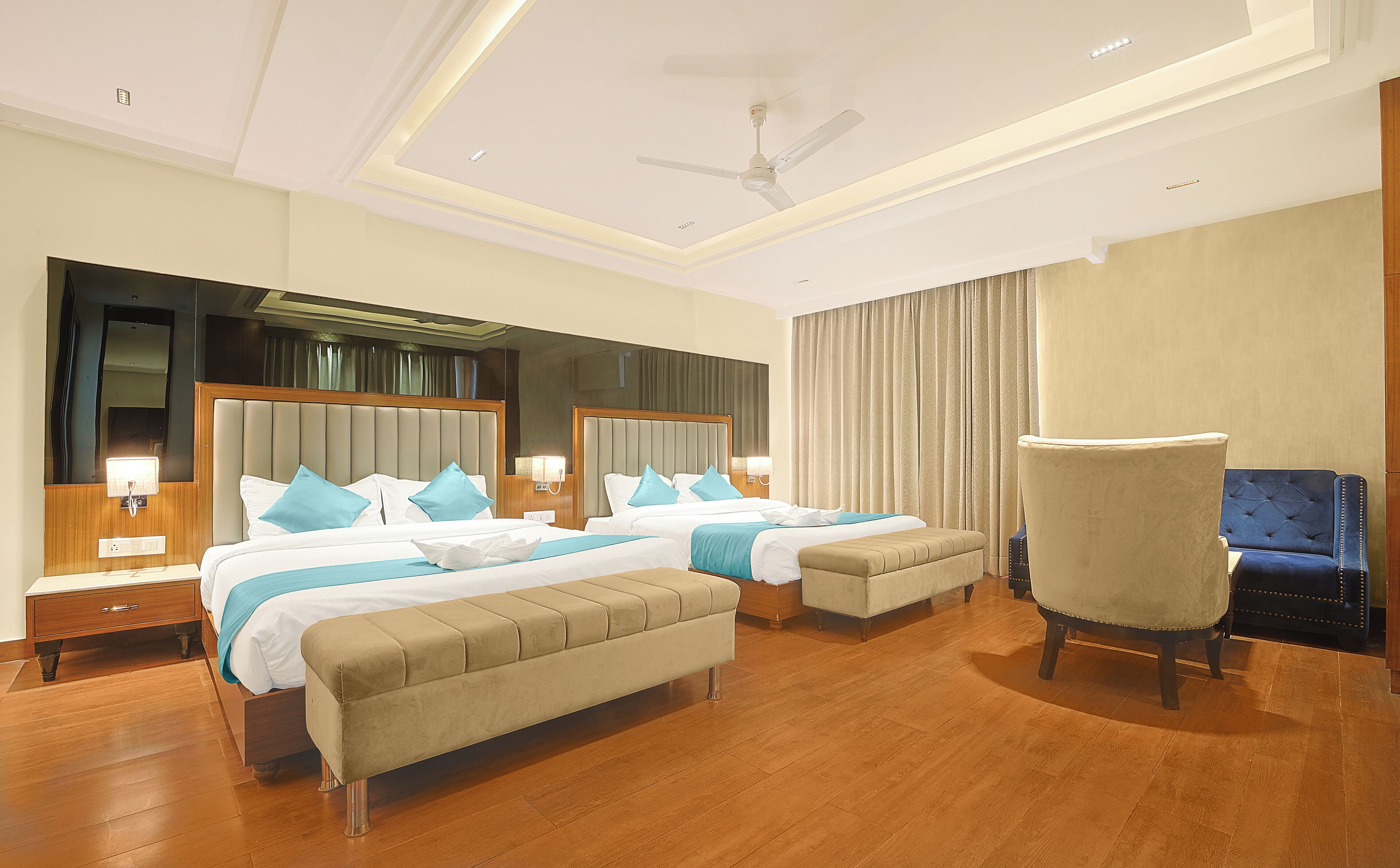 Quarto de hotel com duas camas, um espelho grande, um ventilador de teto e um espaço de estar com uma poltrona.