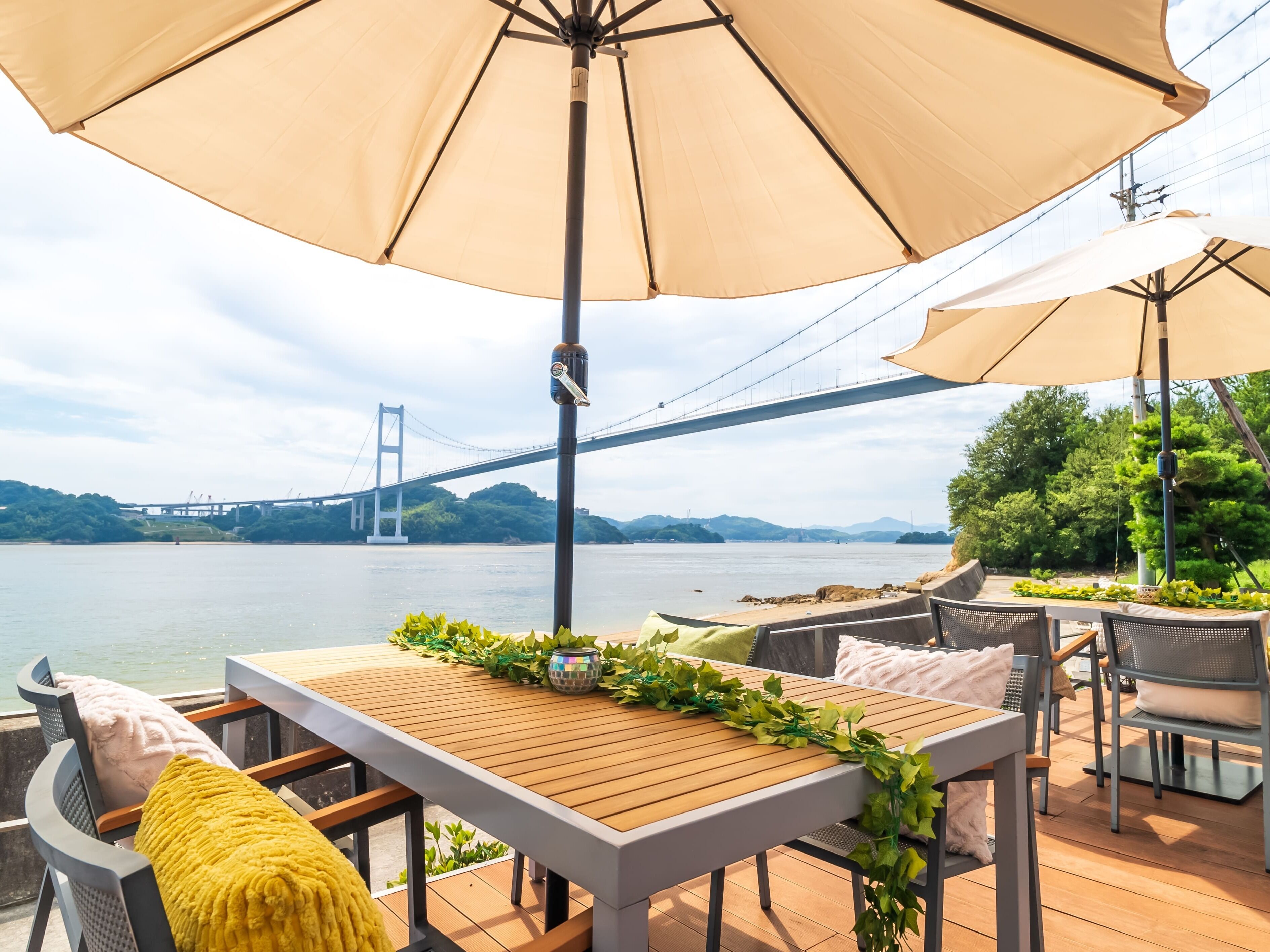 An outdoor seating area with a table and chairs, a large umbrella, and a view of a bridge over water.